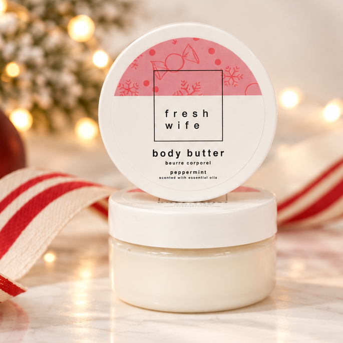 A small white jar of Fresh Wife Body Butter with a round lid leaning on top, styled on a marble surface with holiday decorations including a red ornament, candy canes, a striped ribbon, and warm string lights in the background.