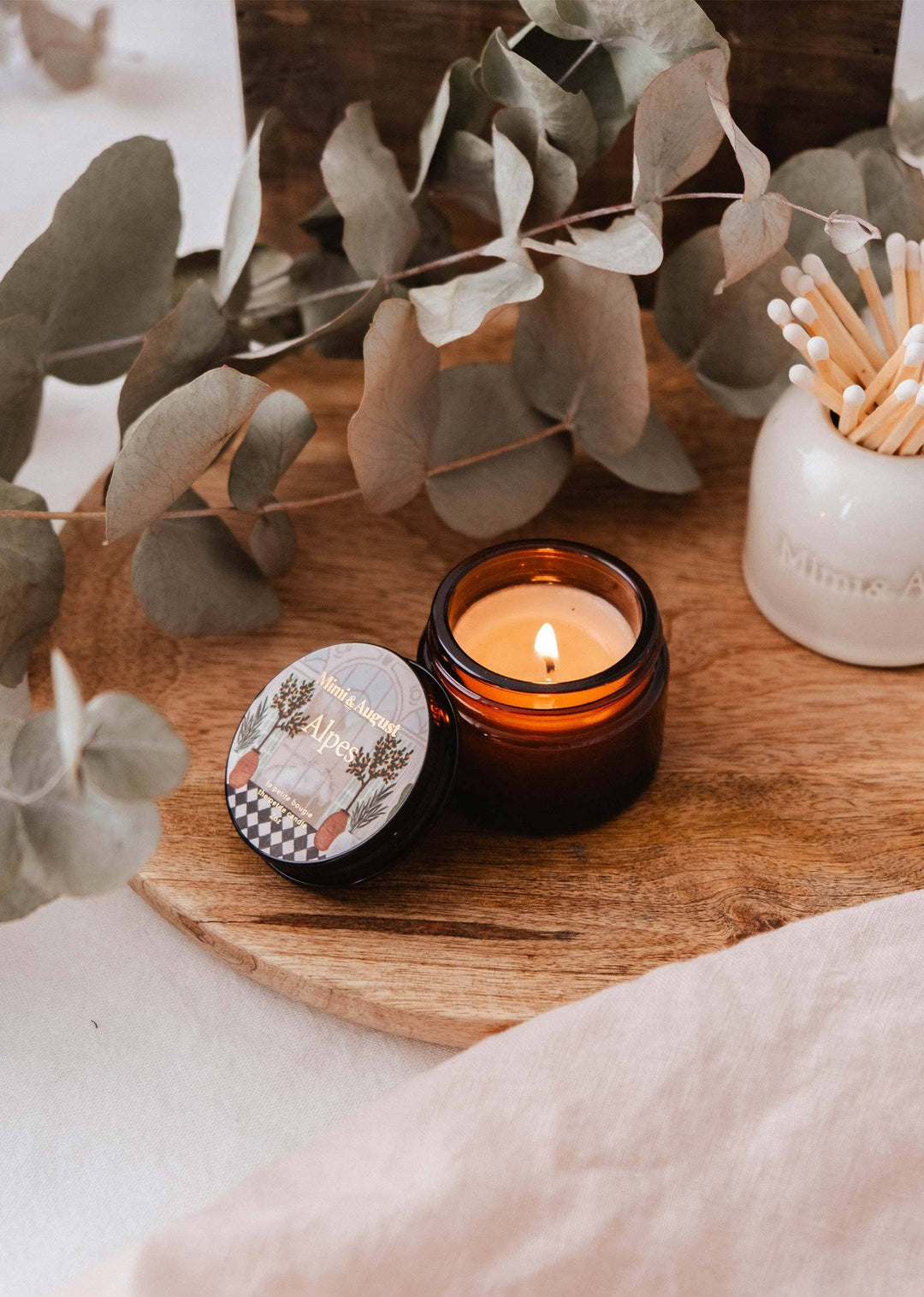 Candle with a decorative lid on a wooden surface with eucalyptus leaves and a match holder.