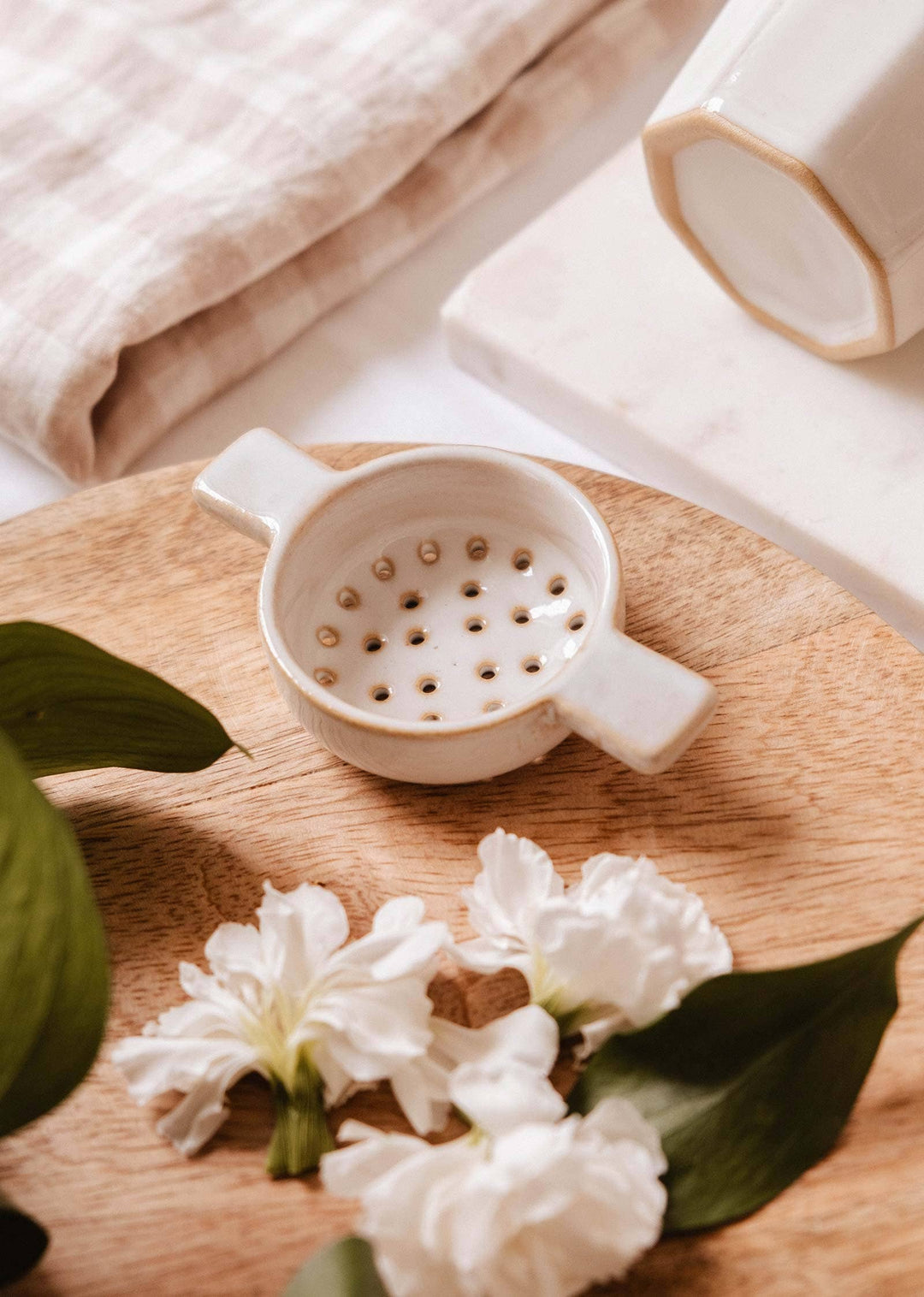 White ceramic strainer on a wooden surface with white flowers and green leaves.