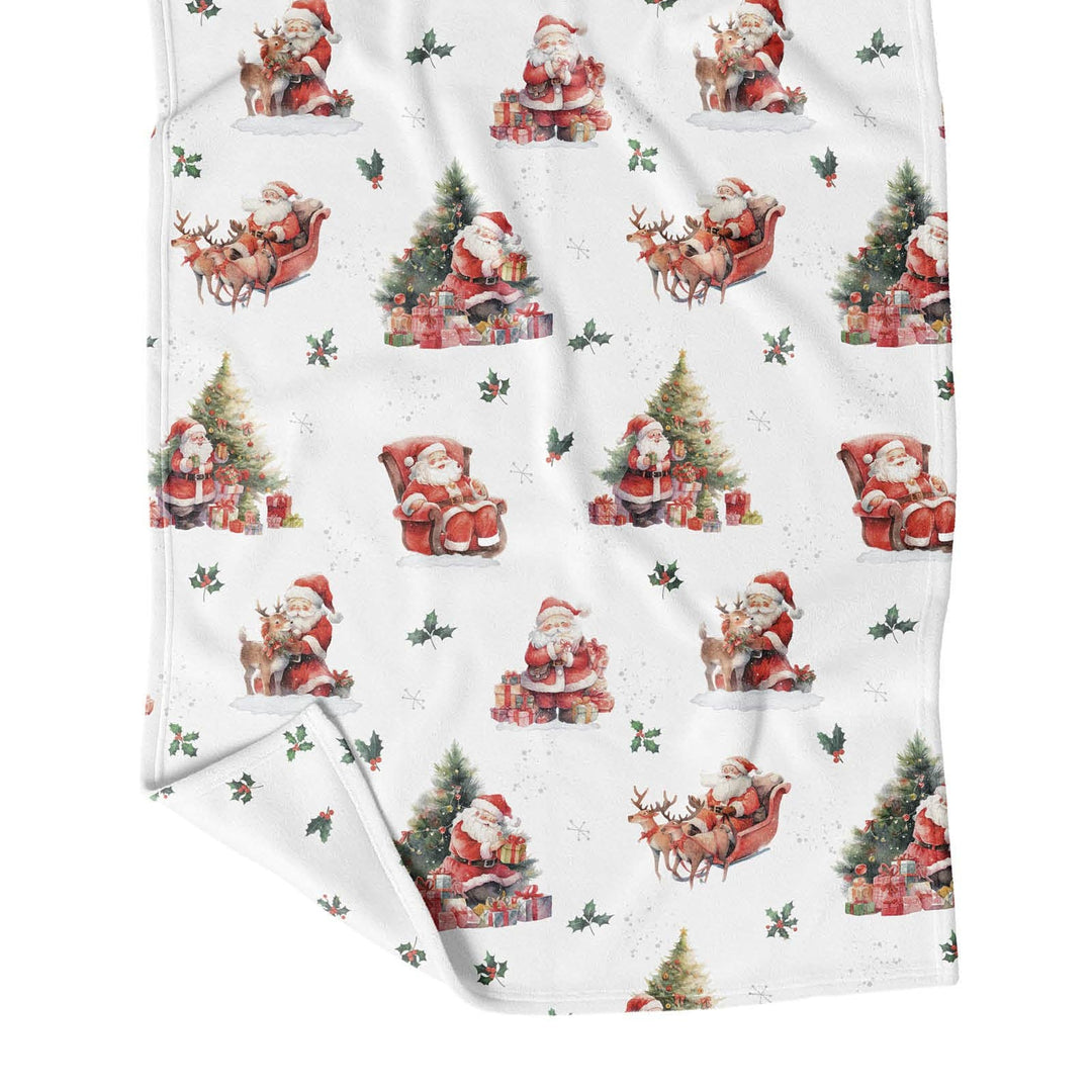 A white fleece blanket featuring a repeating Christmas pattern of Santa Claus in red and green, sitting in sleighs beside Christmas trees and wrapped gifts.