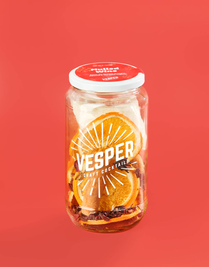A clear glass jar of Vesper Pulled Orange Cocktails with orange slices and spices visible inside, set against a solid coral red background.