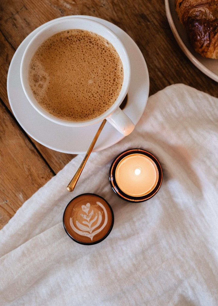 A cozy overhead scene with a white cup of coffee on a saucer, a small lit candle in a dark amber holder, a latte art coffee cup lid, and a white cloth on a wooden table.