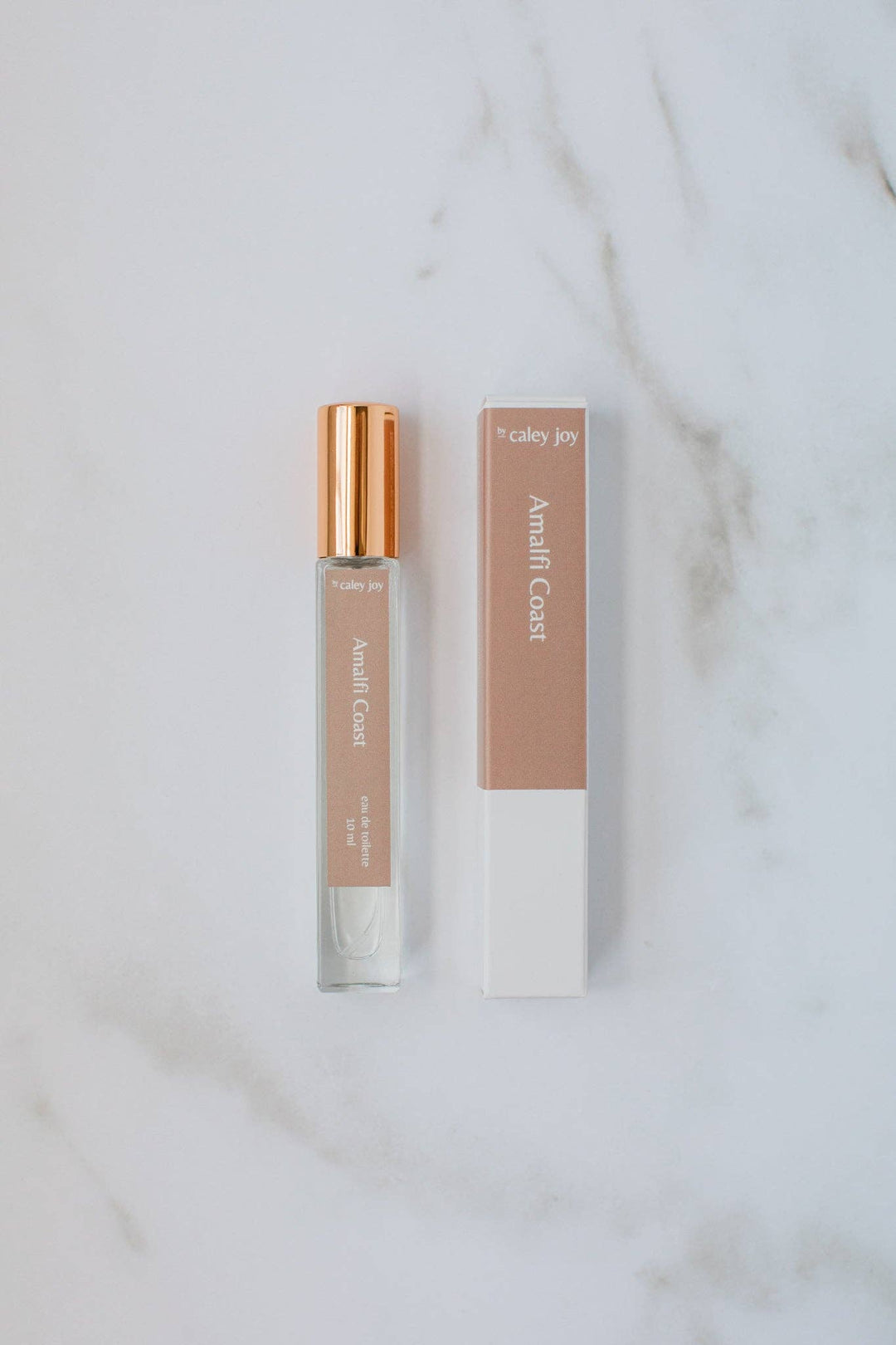 A small clear perfume or fragrance roller bottle with a metallic rose-gold cap stands beside its matching rectangular box on a white marble surface. The packaging is a muted blush pink on the upper half and white on the lower half, with the text &