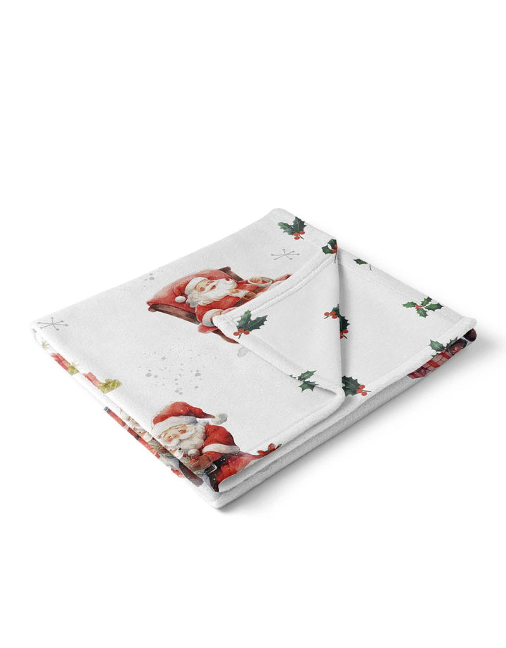 A folded white blanket featuring a Christmas print with Santa Claus, holly leaves, and small snowflakes, shown on a clean white background.