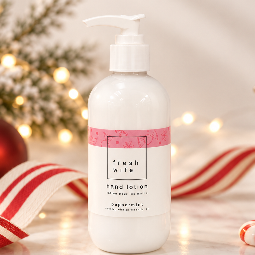 A pump bottle of Fresh Wife Hand Lotion in Peppermint, styled on a marble surface with festive Christmas decorations including a red ornament, candy canes, a striped ribbon, and warm fairy lights in the background.