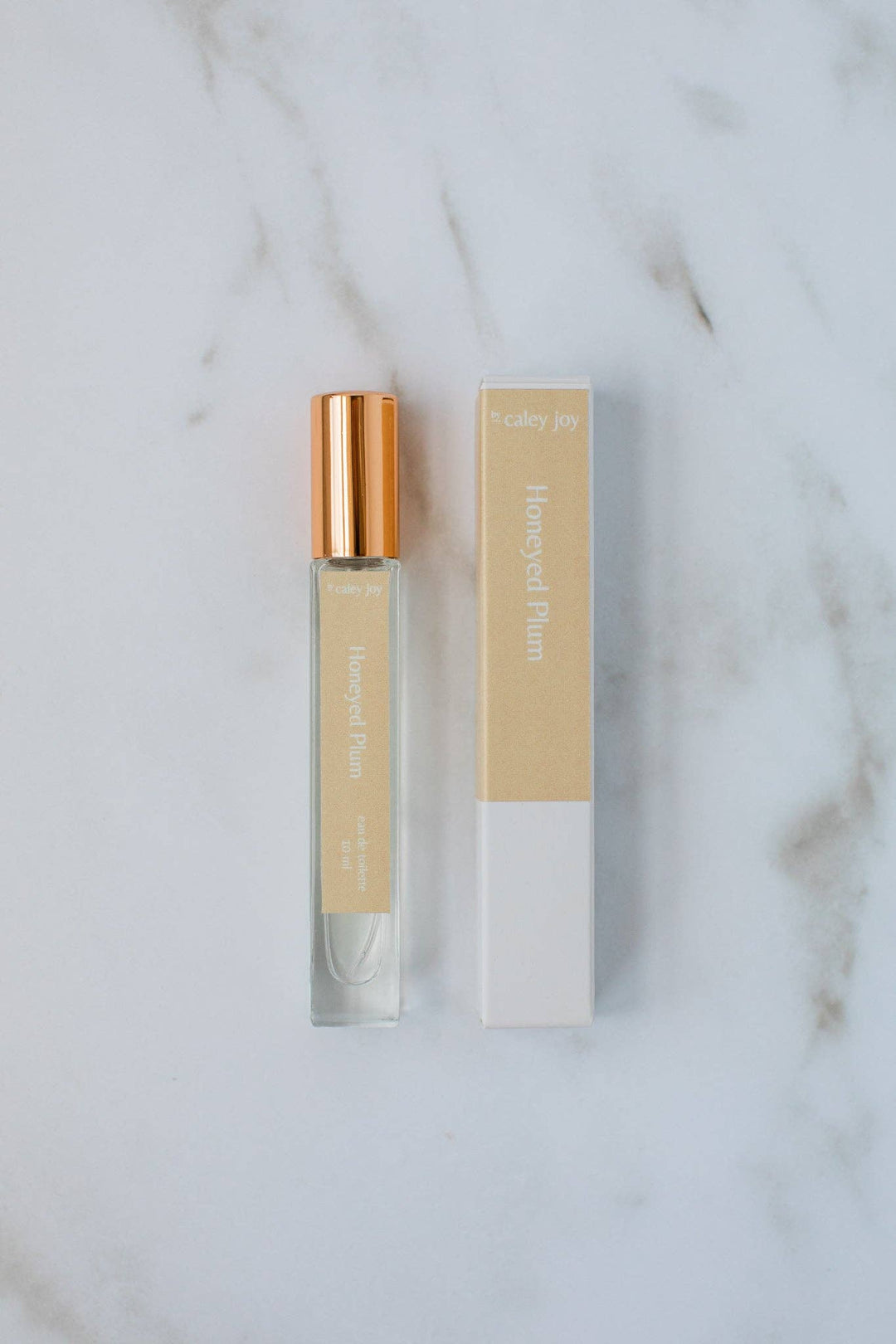 A small clear glass perfume roller bottle with a metallic rose gold cap stands next to its matching beige and white packaging box on a white marble surface.