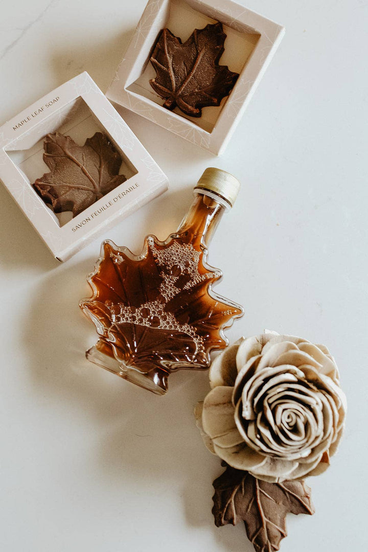 A styled flat-lay of fall-themed bath products on a white surface, including a maple leaf-shaped bottle of amber liquid, two square boxes containing leaf-shaped soaps, and a beige fabric rose with a brown leaf accent.