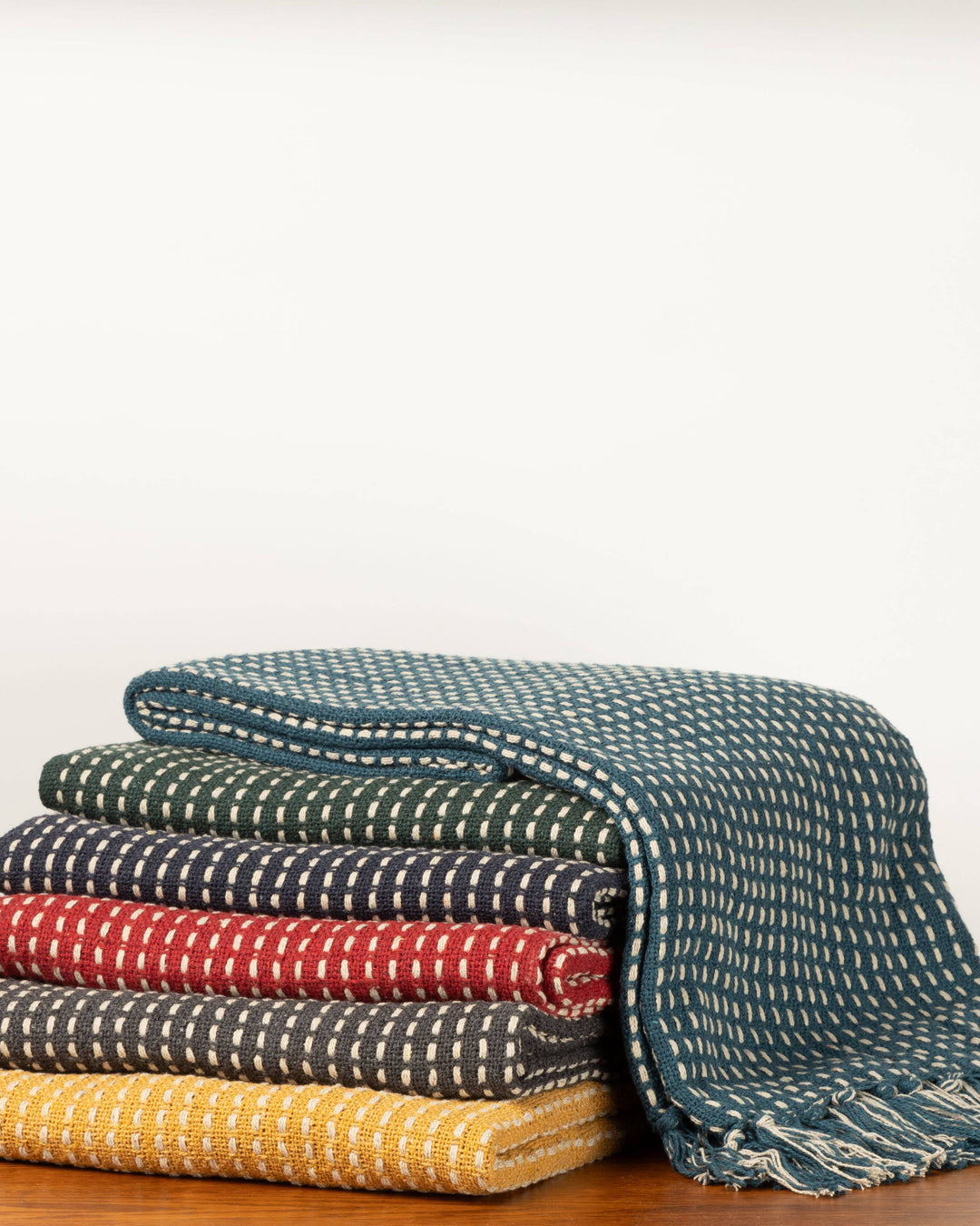 A stack of folded woven throw blankets in teal, green, navy, red, charcoal, and mustard yellow, each with a white dotted pattern, displayed on a wooden surface against a plain white background.