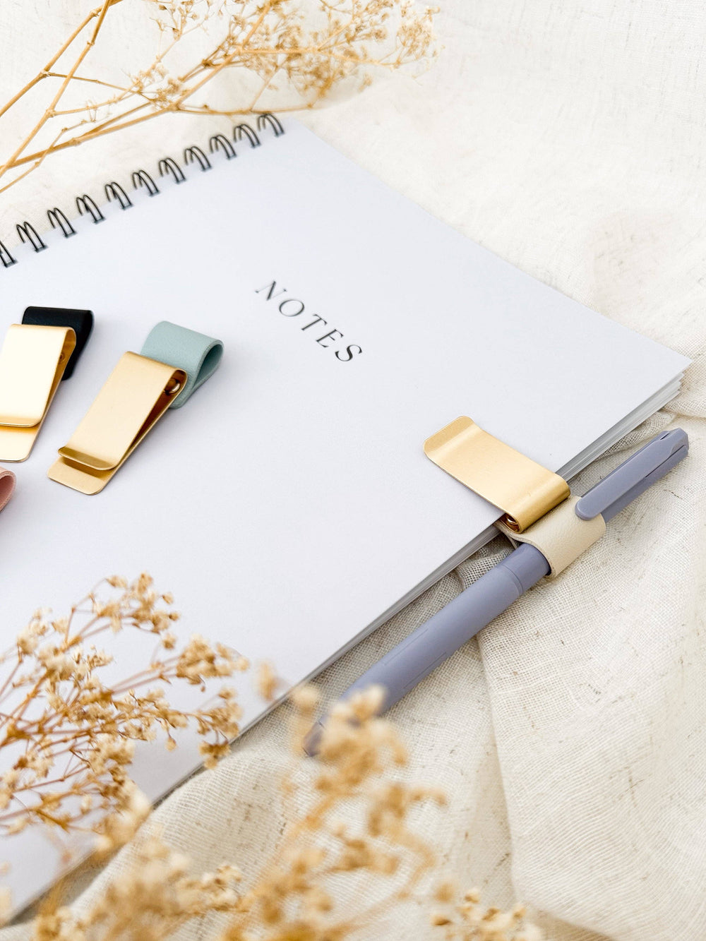 Notebook with a pen and decorative clips on a light surface with dried flowers.