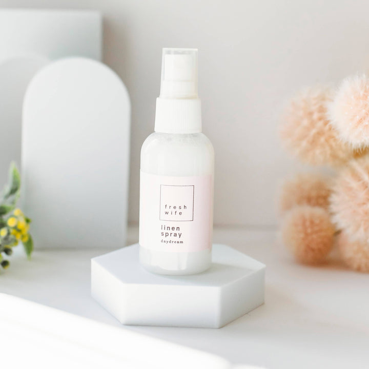 A white bottle of Daydream Linen Spray with a spray pump, displayed on a white platform with a blurred background featuring a decorative object and some flowers.