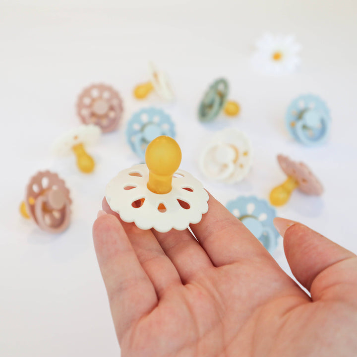 Hand holding a white pacifier with colorful pacifiers in the background on a light surface.