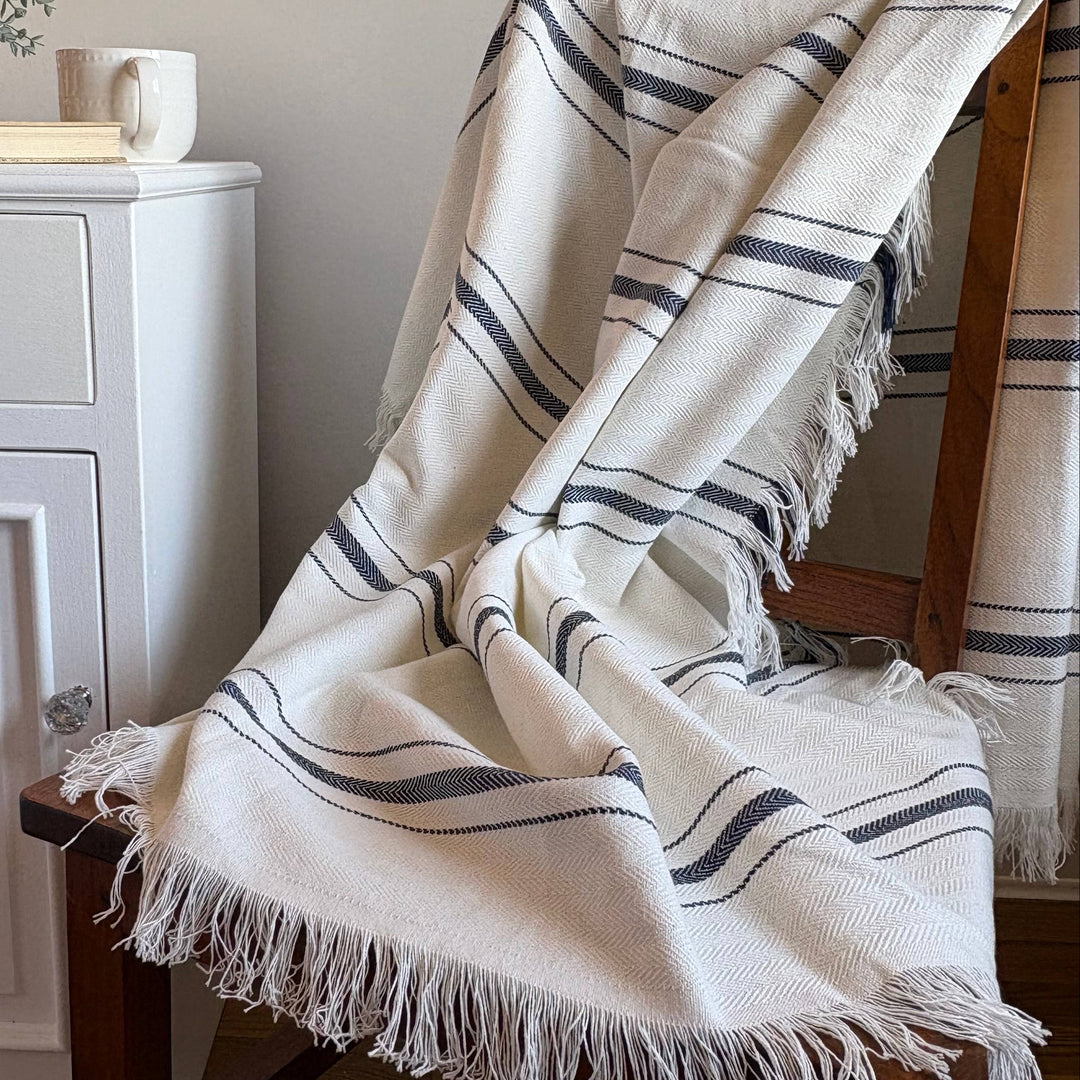 A cream-colored woven throw blanket with dark navy stripes and fringed edges draped over a wooden chair in a cozy room setting.