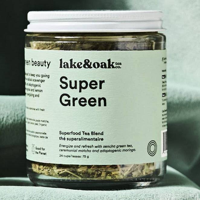 Jar of 'lake & roo' Super Green on a green cloth with a glass of tea, lemon, and spoon.