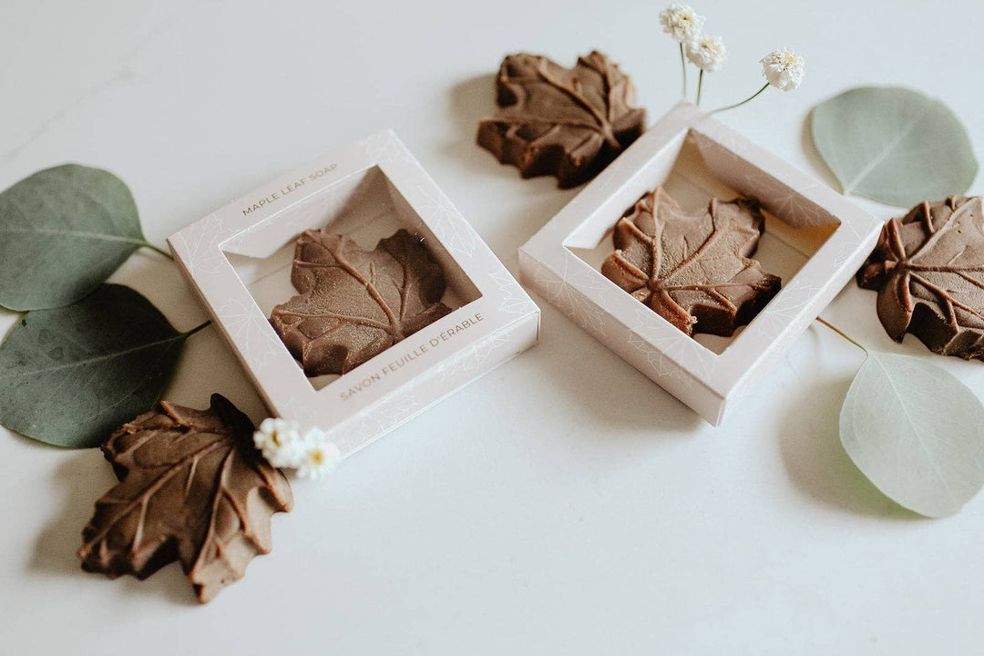 Two square light wood boxes containing brown leaf-shaped soaps, arranged on a white surface with eucalyptus leaves and small white flowers.