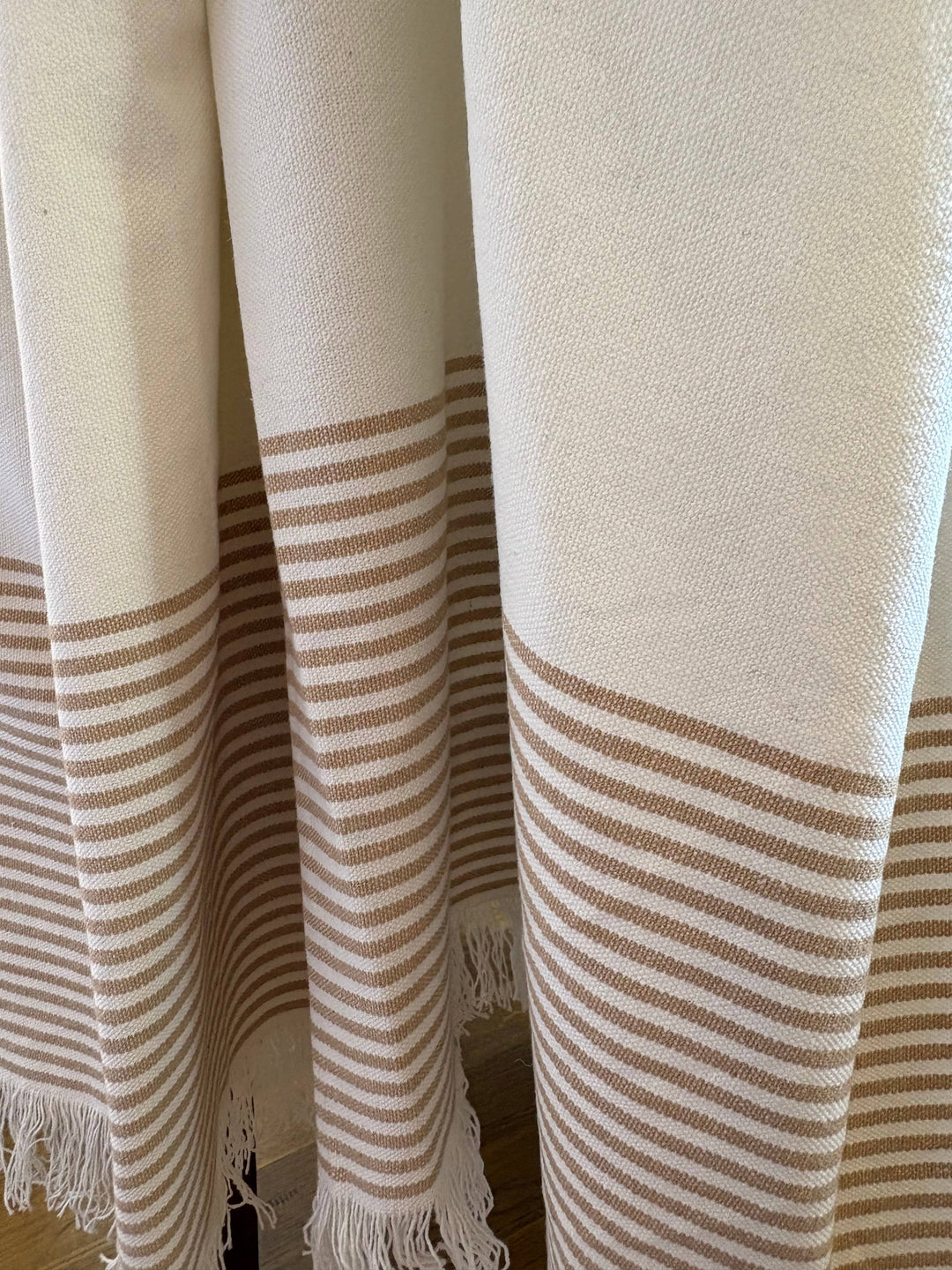 Close-up of a woven textile with cream and tan stripes and fringe, likely a throw blanket or curtain panel.
