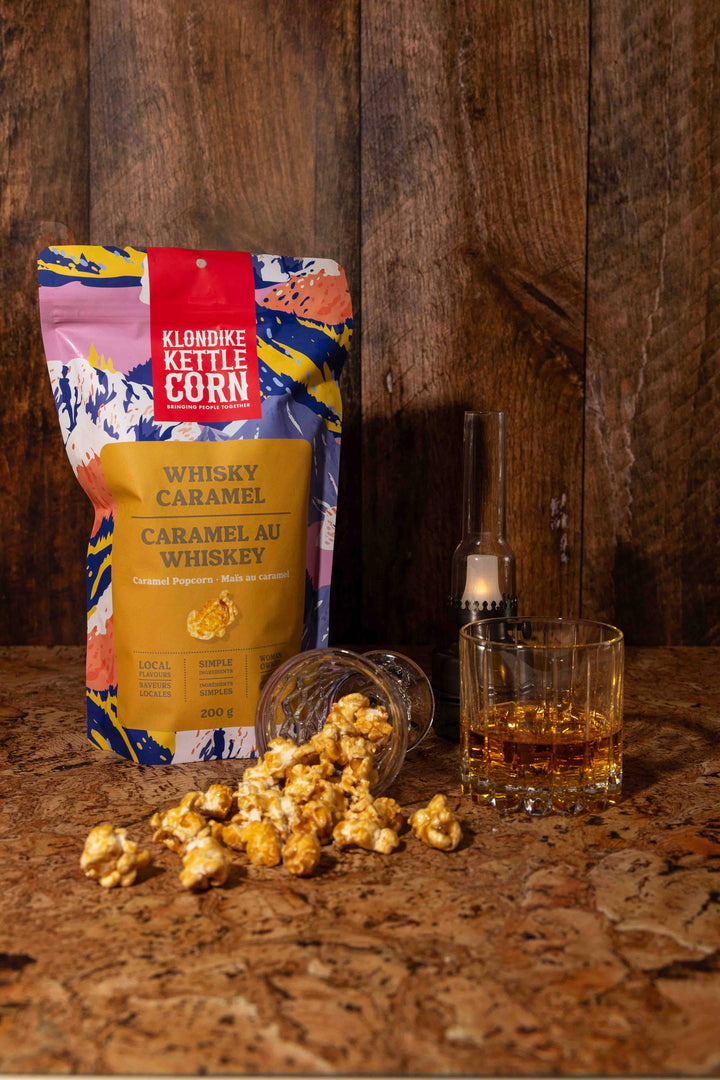 Klondike Kettle Corn bag with popcorn and a glass of whiskey on a wooden surface