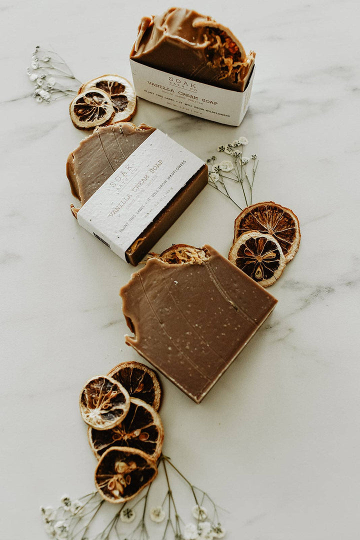 Three bars of soap with a white box on a marble surface with dried oranges and flowers.