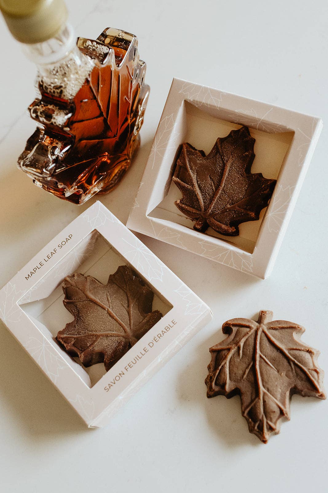 Three maple leaf-shaped soaps displayed in white square boxes, with one soap outside the box on a light surface. A small amber glass bottle with a gold cap is partially visible in the upper left.