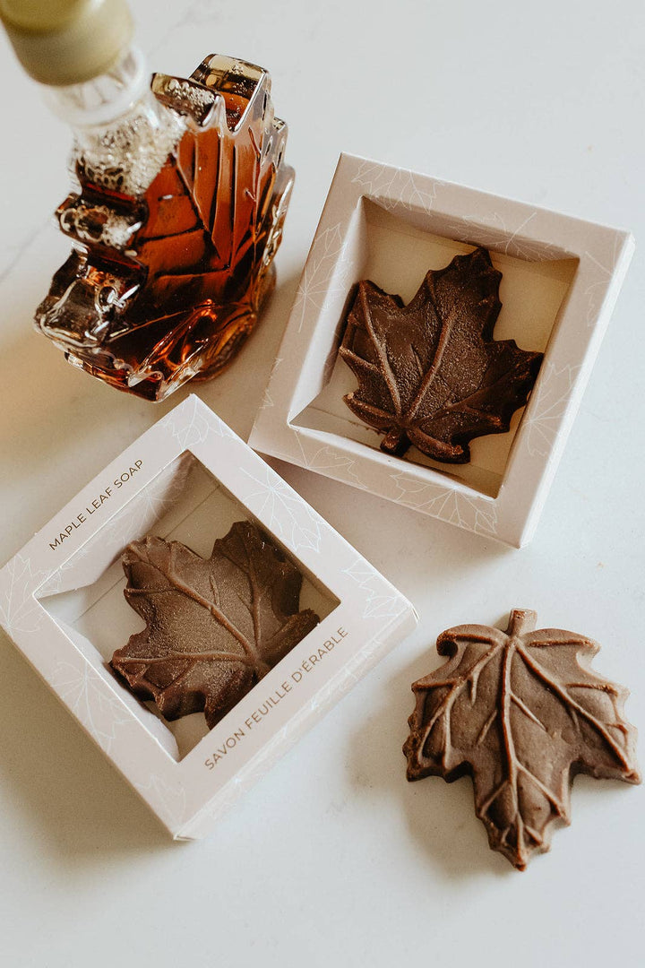 Three maple leaf-shaped soaps displayed in white square boxes, with one soap outside the box on a light surface. A small amber glass bottle with a gold cap is partially visible in the upper left.