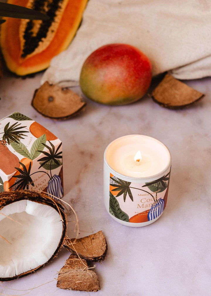 Candle with coconut and mango design surrounded by coconuts and a mango on a light background