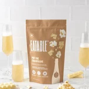 A 125g bag of Eatable Pop The Champagne Popcorn, with images of popcorn and a champagne bottle on the front, displayed alongside two glasses of champagne and a bowl of popcorn.